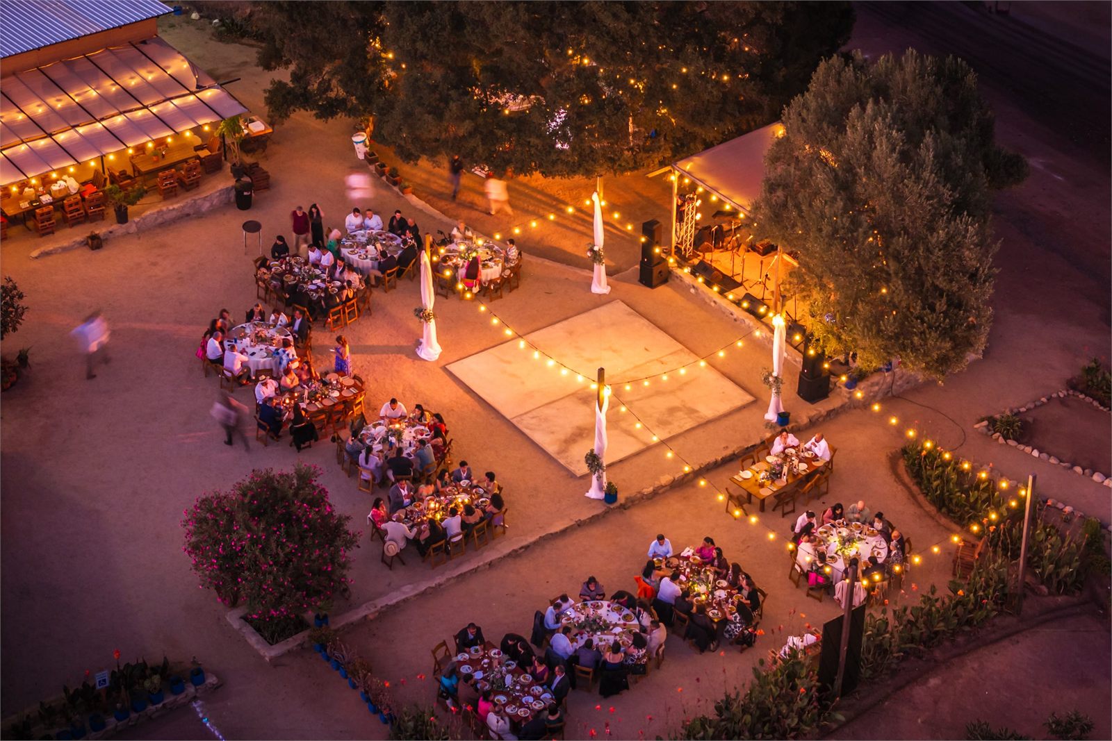 An evening event seen from above, tables and a dance floor lit by warm string lights