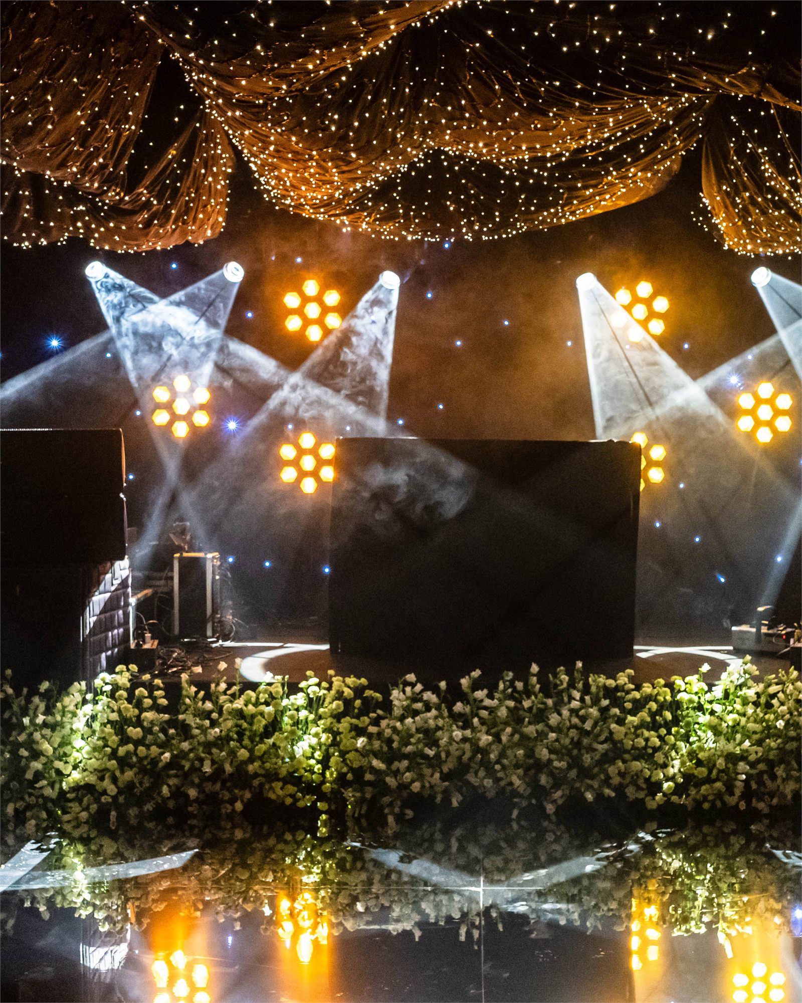 A stage under crossed spotlights banked with white flowers beneath a fairy-light canopy