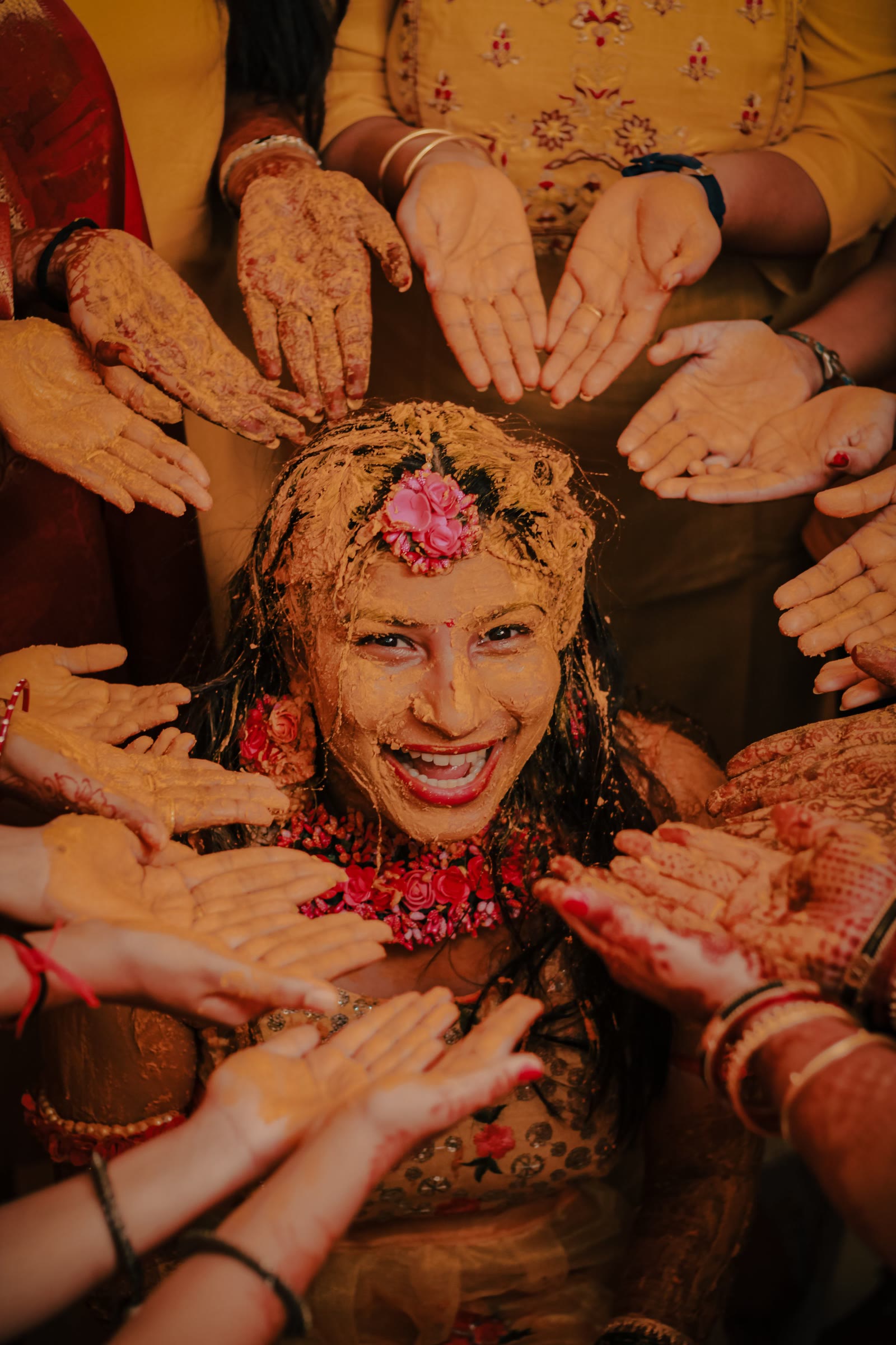 A bride covered in turmeric, laughing, surrounded by the hands of her family