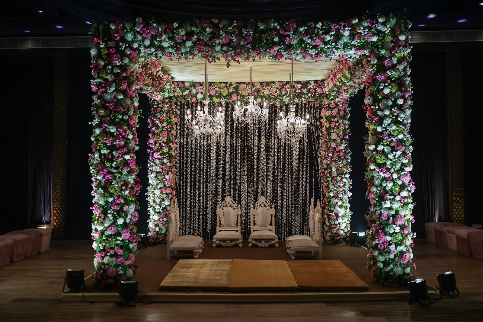An indoor mandap framed in pink and white florals beneath crystal chandeliers