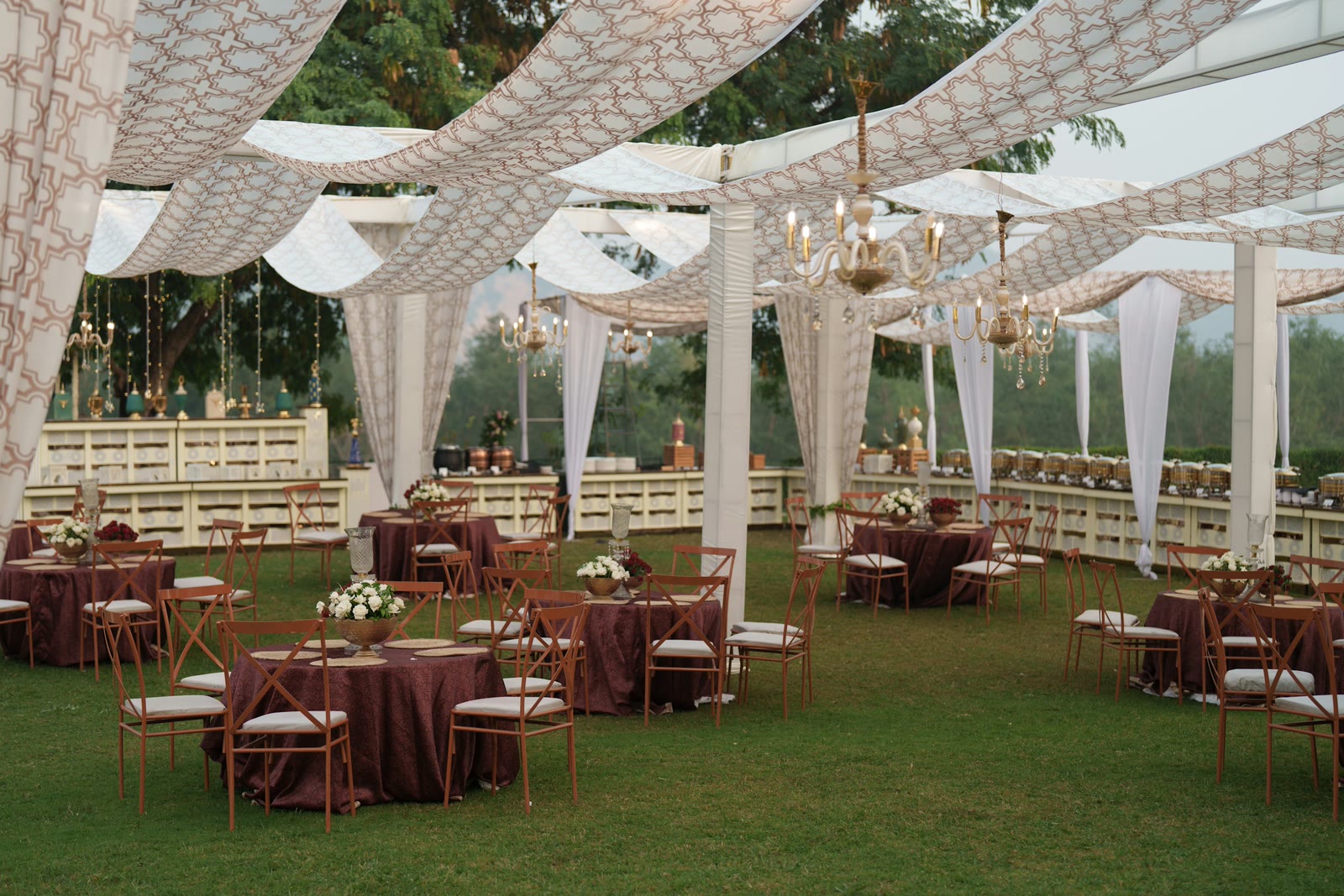 An outdoor reception laid with round tables under a draped canopy and chandeliers